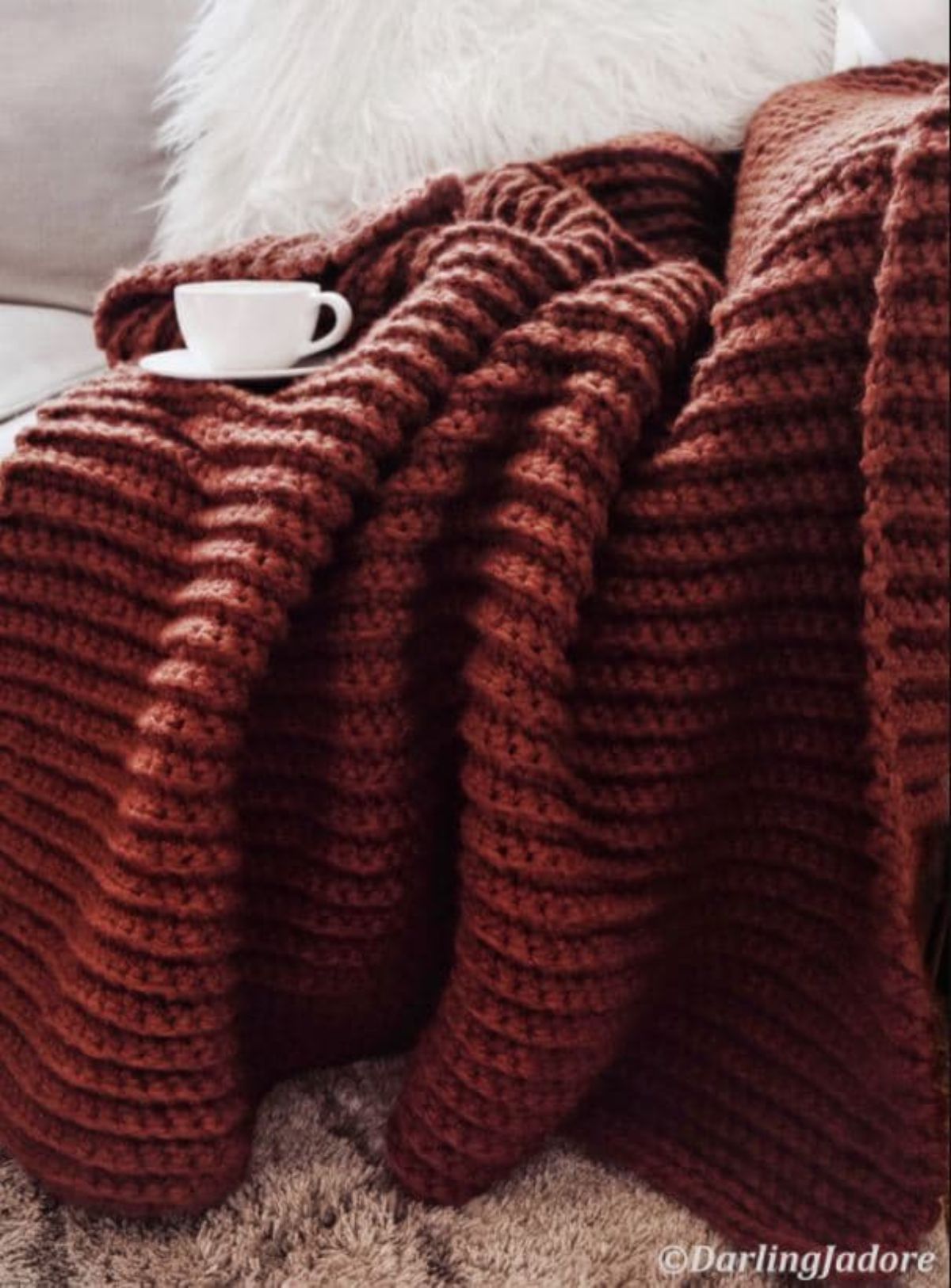 Dark orange chunky crochet blanket draped over a cream sofa with a white cup and saucer balanced on the blanket.