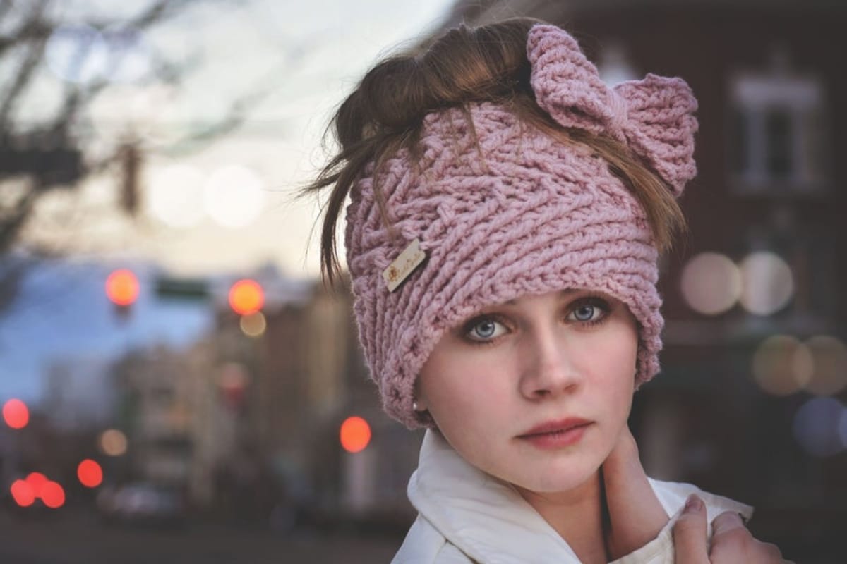 A brunette woman standing outside wearing a pink crochet hat with a messy bun pulled through and a large pink bow on the side.