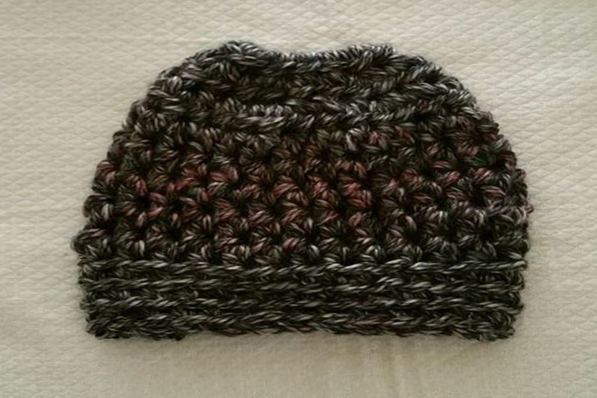 Black crochet hat with some pink and cream speckles and a hole at the top on a cream background.