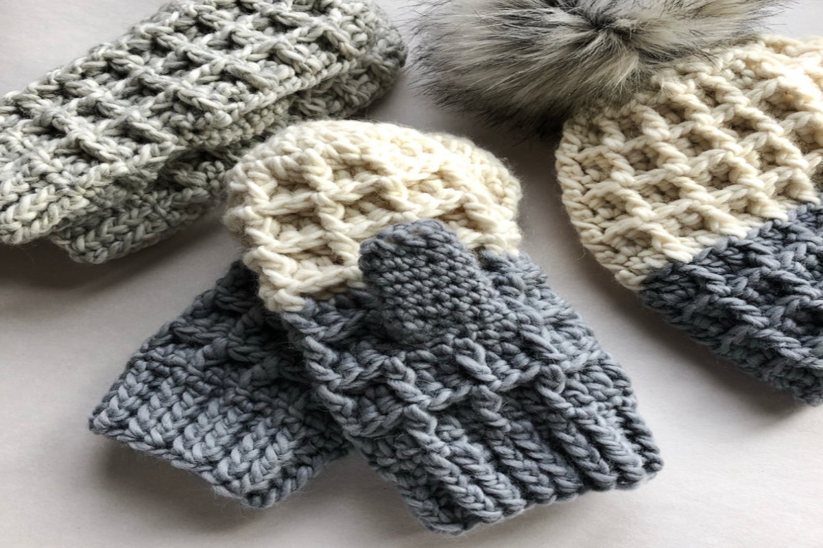 Waffle stitched cream and gray chunky mittens with a hat in the same colors with a fluffy bobble on top laid on a beige table.