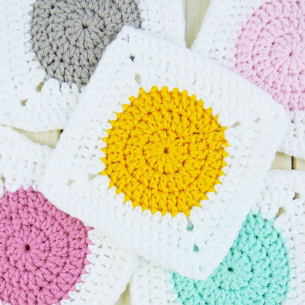  White granny square with a mustard yellow circle in the center on top of pink, green, and gray squares with the same design.