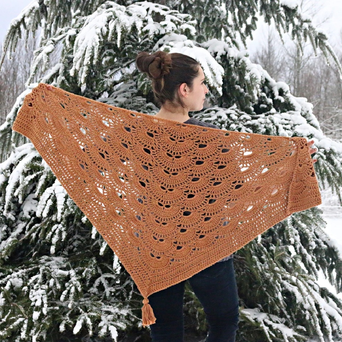 Brunette woman standing by a snowy tree with an orange crochet shawl and a large orange tassel hanging at the bottom.