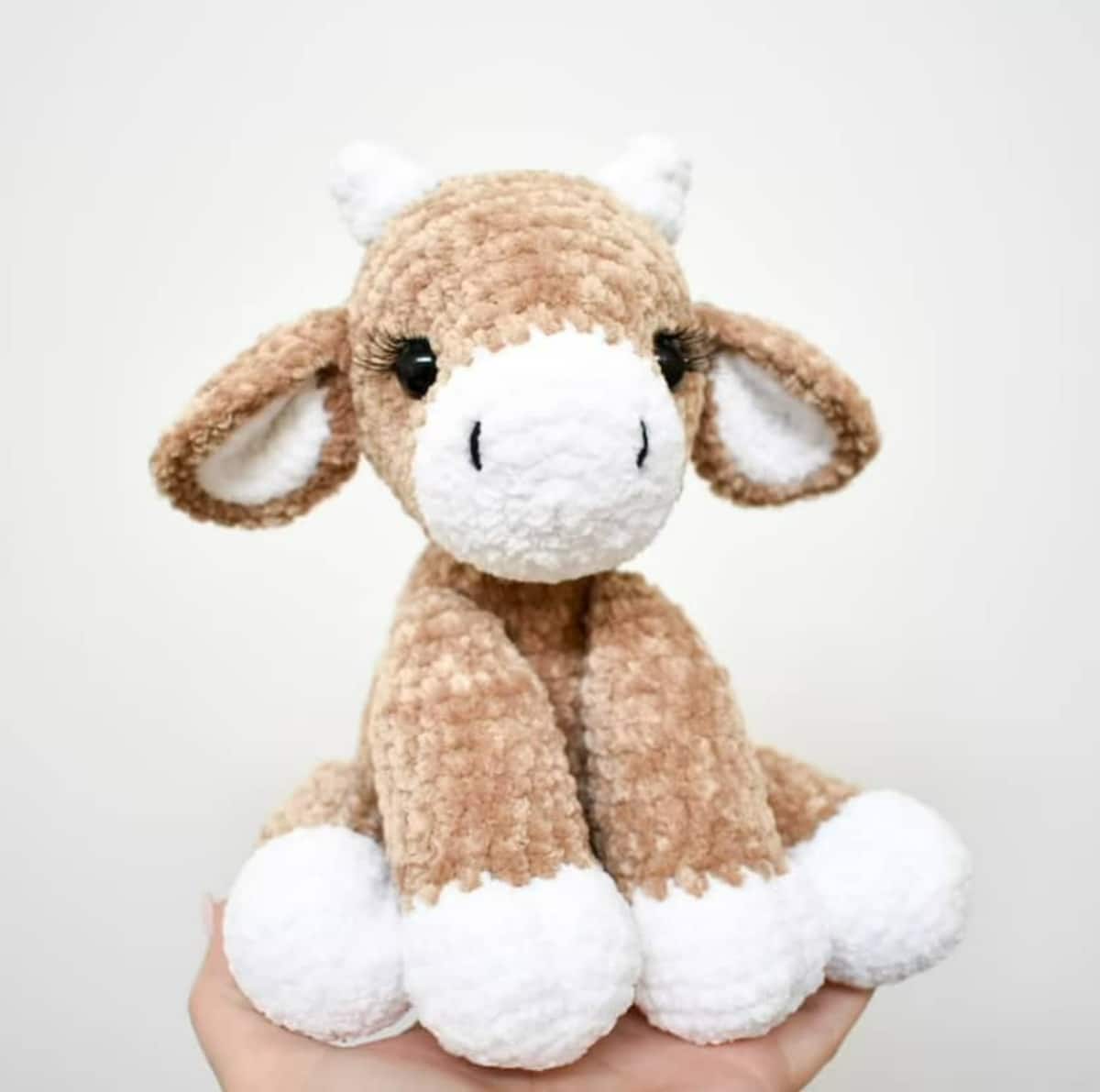 A hand holding a brown crochet cow with white feet, ears, and horns sitting down with its back legs spread behind them.