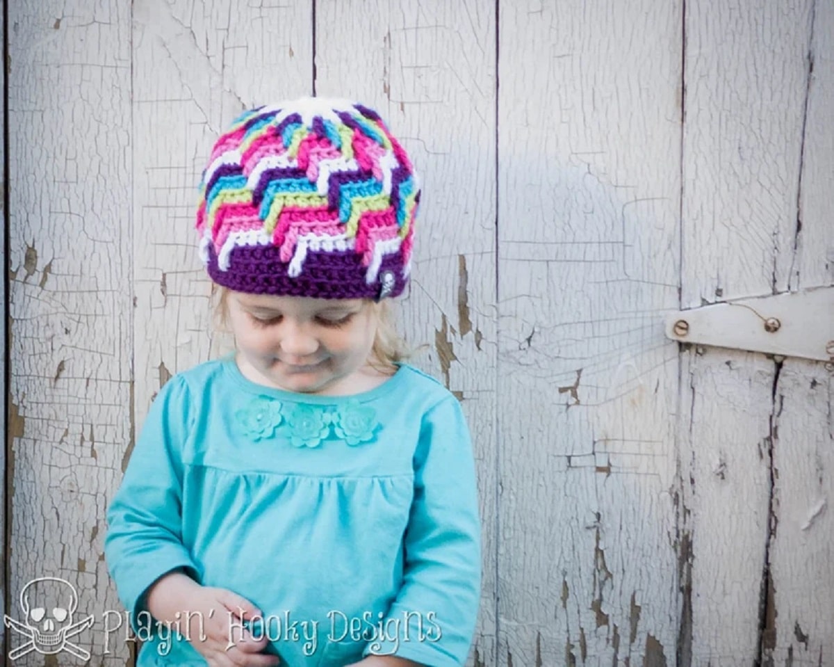 Young child in blue wearing a purple crochet hat with a white, pink, purple, yellow, and blue zig-zag pattern on top.