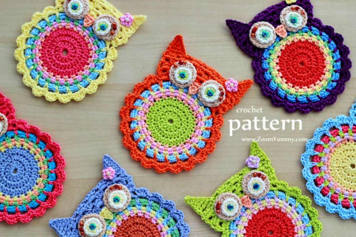  Seven brightly colored owl crochet coasters with white eyes and pointed ears scattered on a pale wooden table.