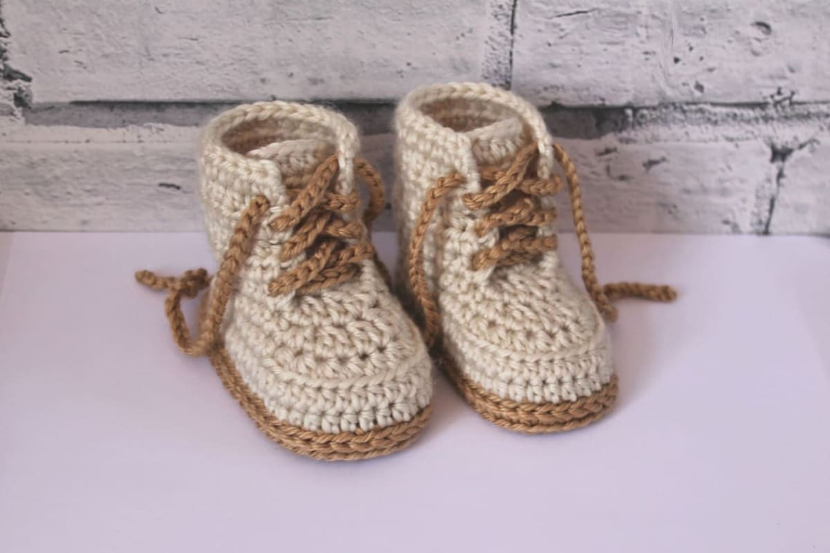 Beige high-top combat style crochet baby booties with brown laces to secure and matching brown soles on a white table.