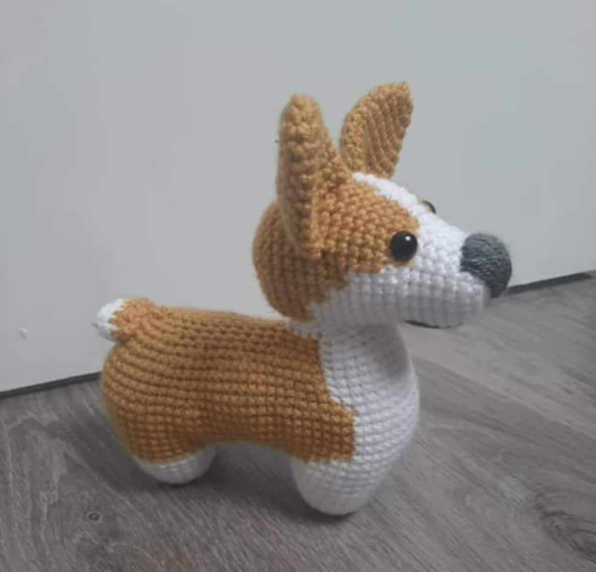 Crochet stuffed Corgi facing sideways with a brown back and ears and a white face and body standing on a wooden floor.