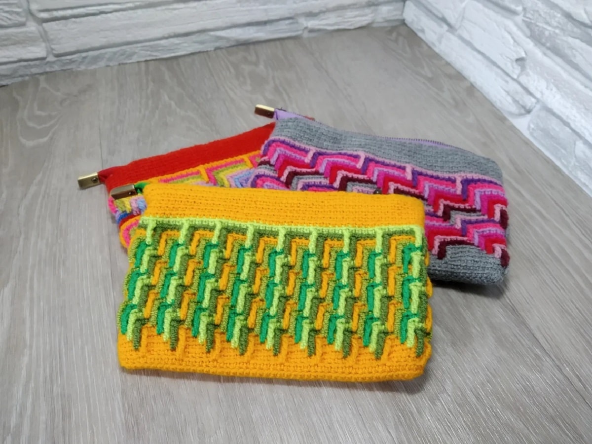 An orange, yellow, and green crochet cosmetic bag lying on top of a gray, purple and pink bag, and a red and orange bag.
