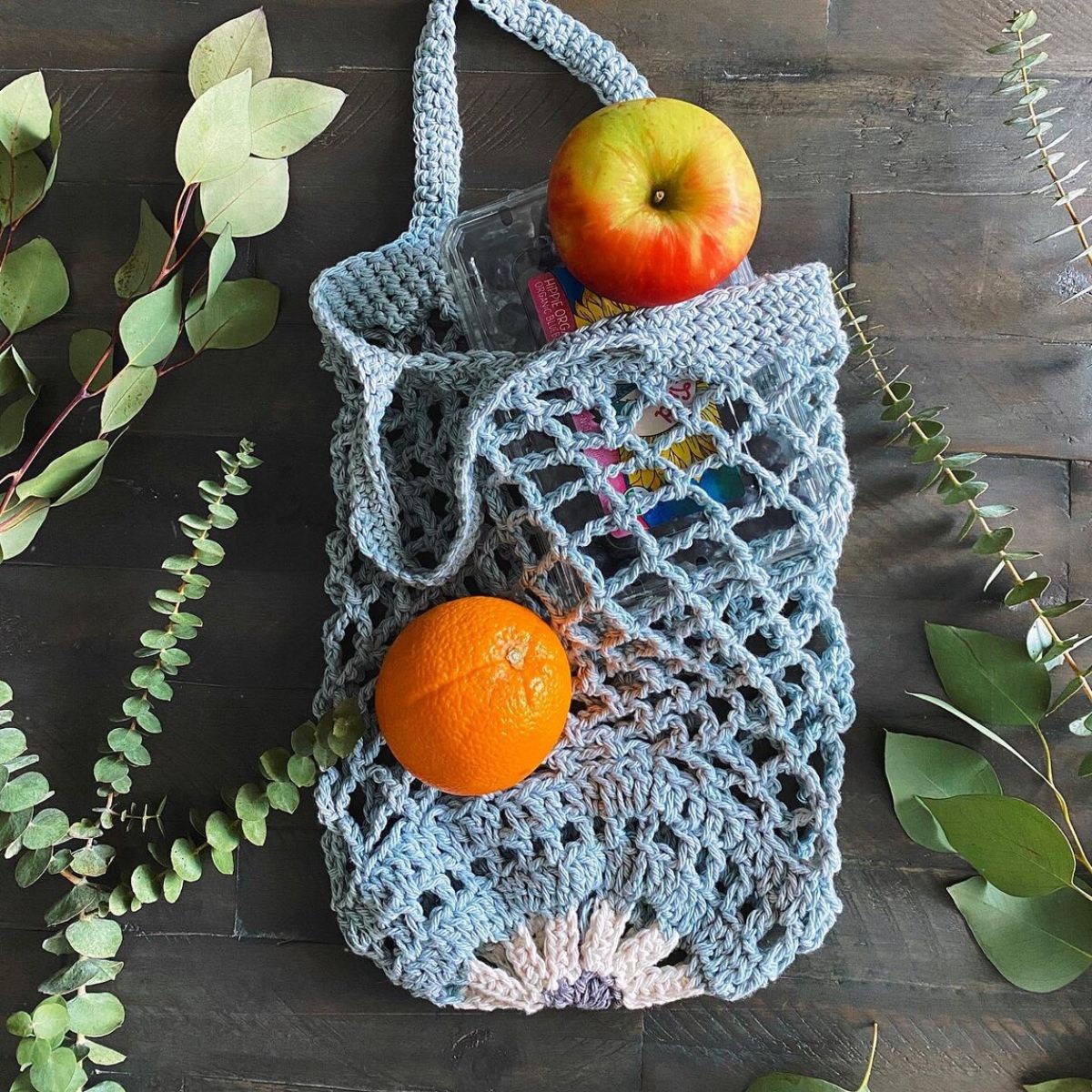 Pale blue crochet market bag with a white flower at the bottom with oranges and apples coming out of the bag and two short handles.