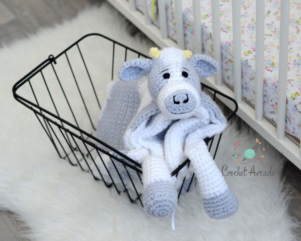 A white and pale blue crochet blanket with a stuffed baby cow’s face with black nostrils and eyes and cow legs, folded up in a black basket.