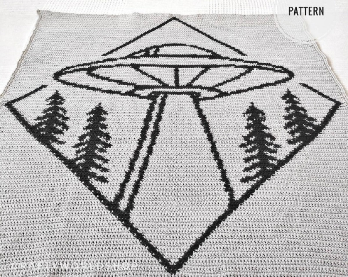 Large light gray wall hanging with a black diamond in the center with a UFO and trees inside it.