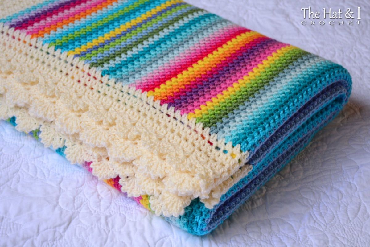 A folded rainbow striped crochet blanket with a cream lace trim along the bottom on a white background.