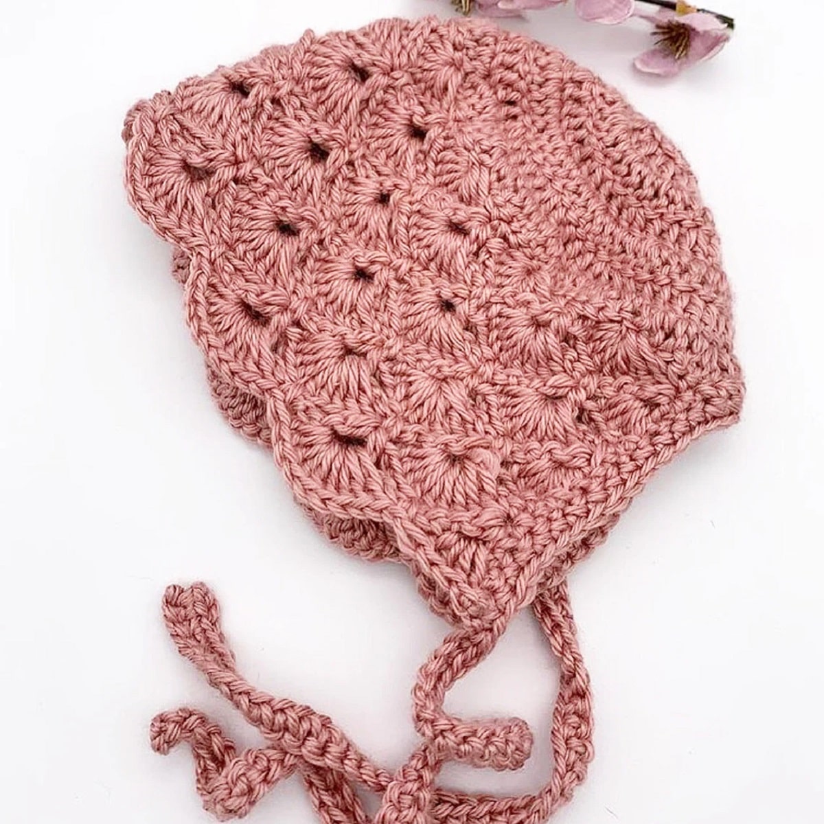 Pink crochet baby bonnet with fan like stitching and a scalloped edge with braided string to secure the bonnet on a white background.