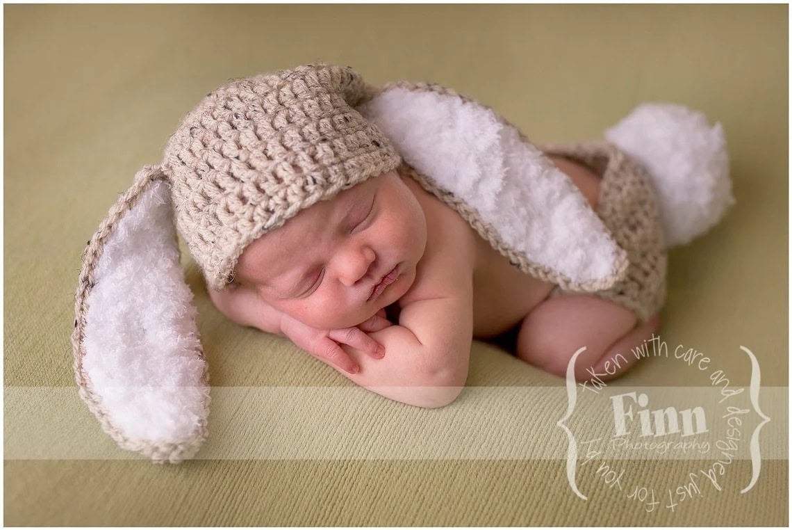 A newborn baby asleep wearing a beige crochet hat with long white bunny ears and a beige diaper cover with a white bunny tail.