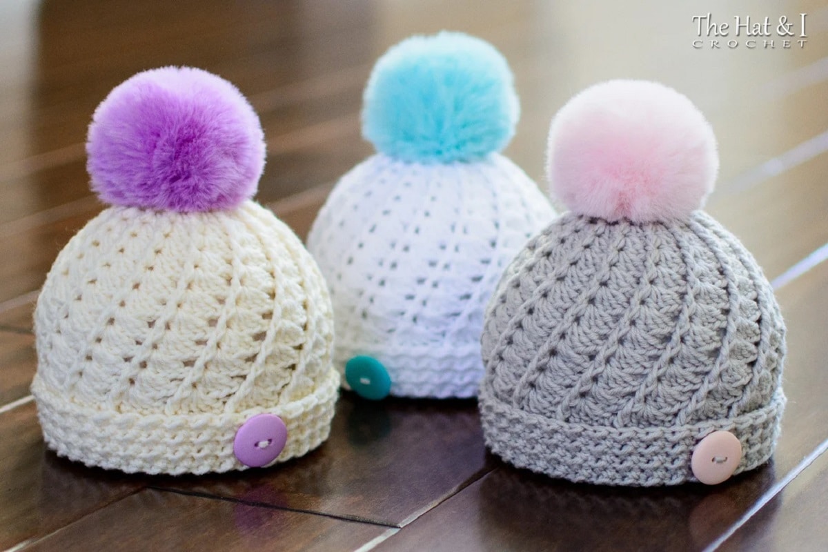A grey crochet bonnet with pink bobble on top, a white bonnet with blue bobble, and cream bonnet with purple bobble standing on a wooden floor.