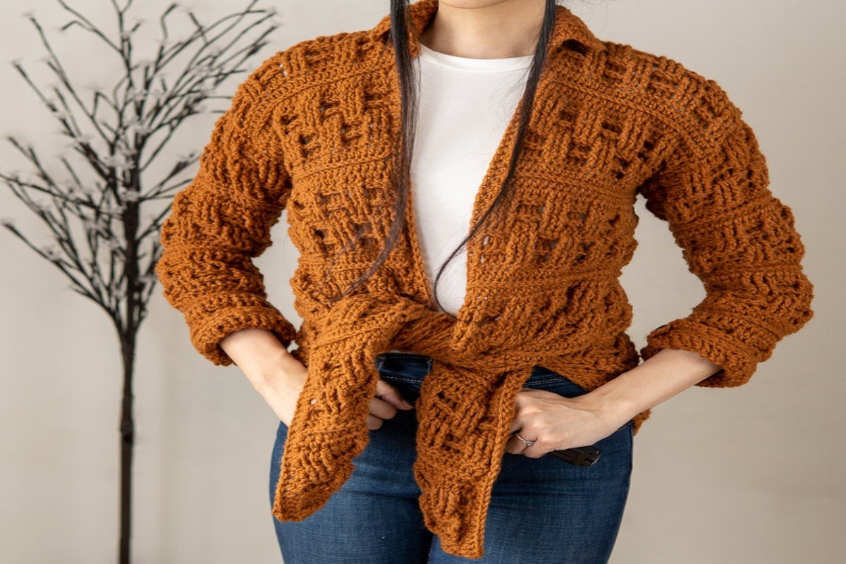 Woman wearing a rust colored chunky crochet cardigan with it tied into a knot at her hips and the sleeves rolled up.
