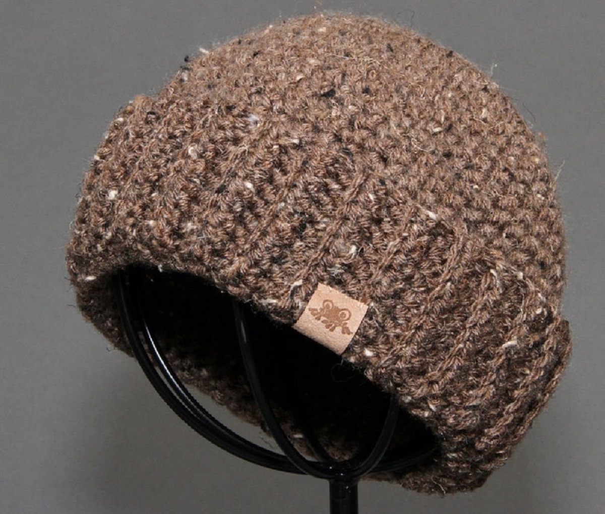 Brown and white speckled crochet beanie sitting on a black metal display unit with a gray background.