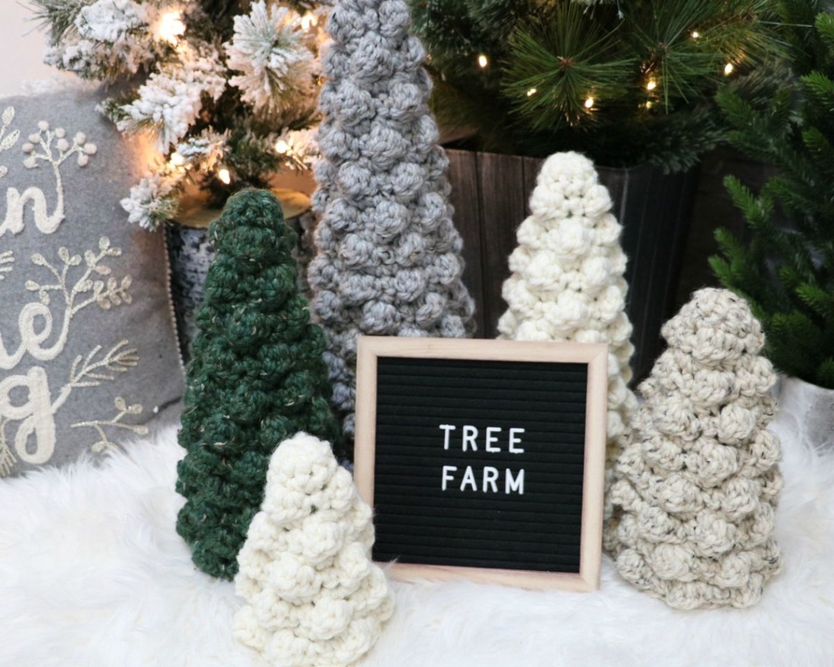 A small white crochet Christmas tree next to larger trees in gray, white, and green and a blackboard sign that says tree farm.