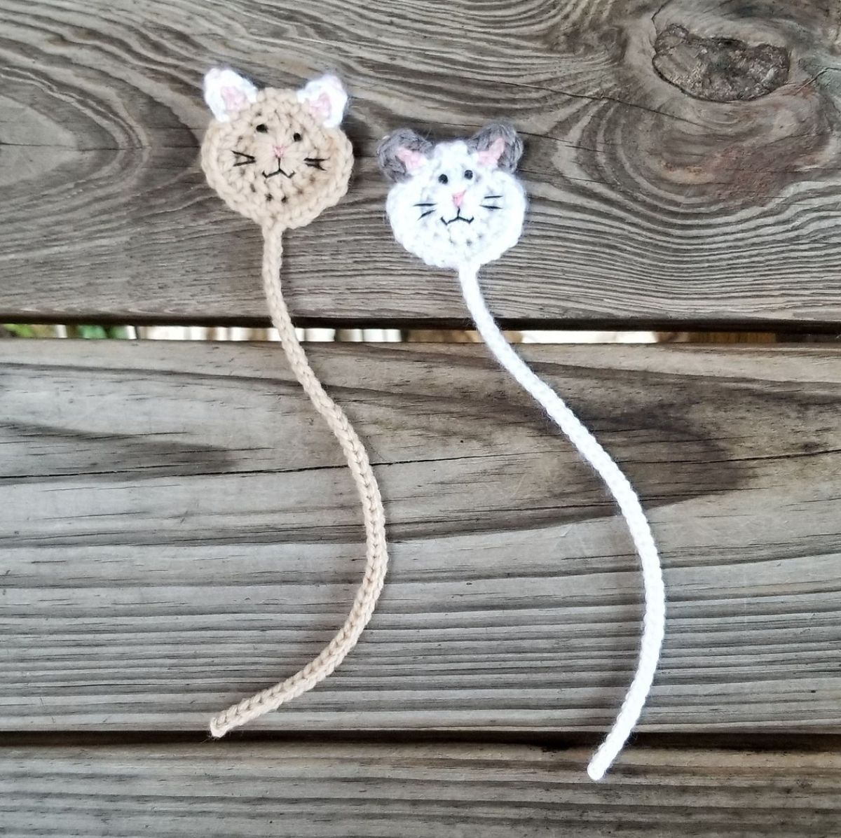 White and beige crochet cat faces with black stitching and thin stitched bodies to use as bookmarks on a gray wooden table. 