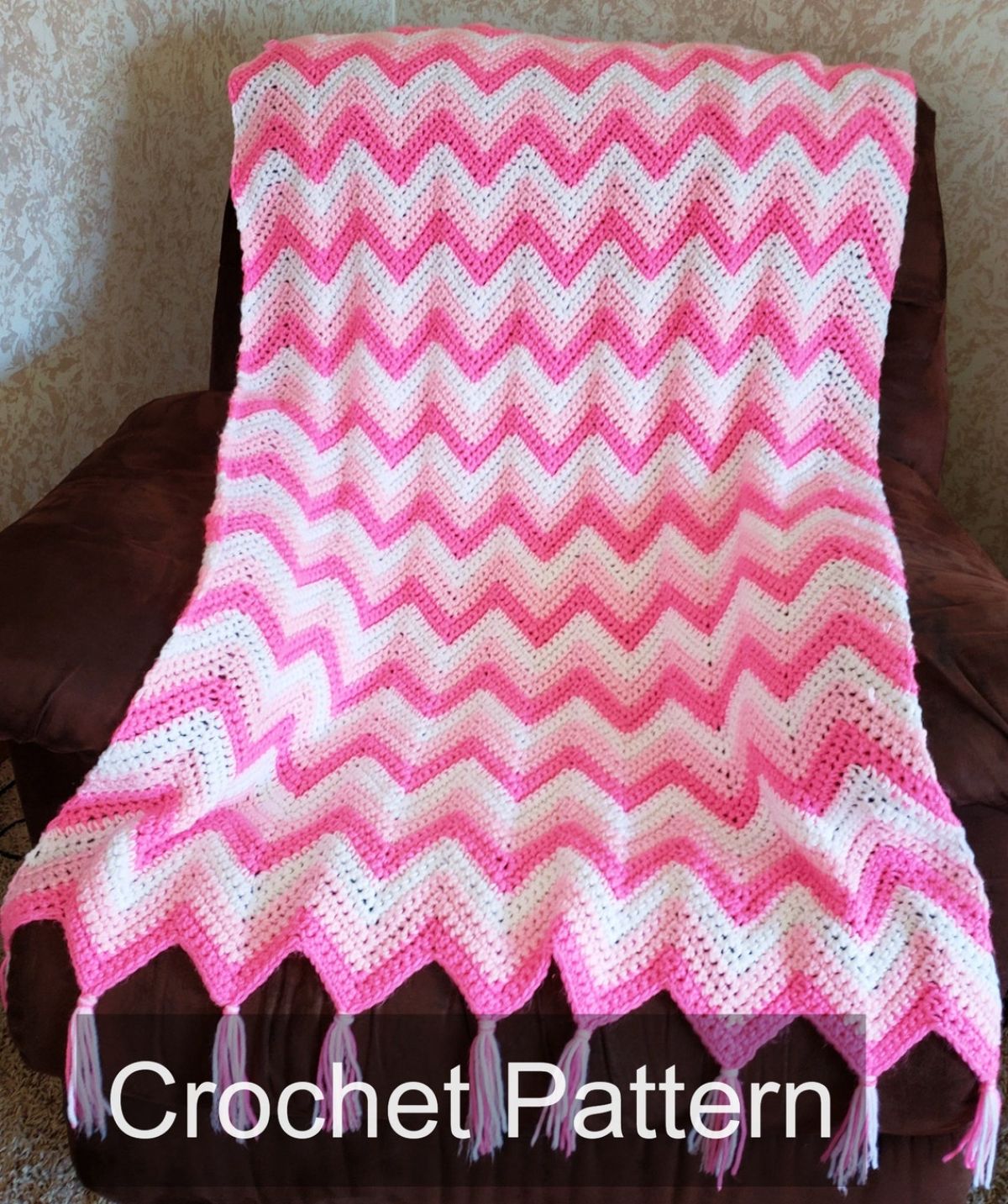 Light pink, pink, and white crochet chevron style blanket with pink tassels on the bottom draped over a brown chair.