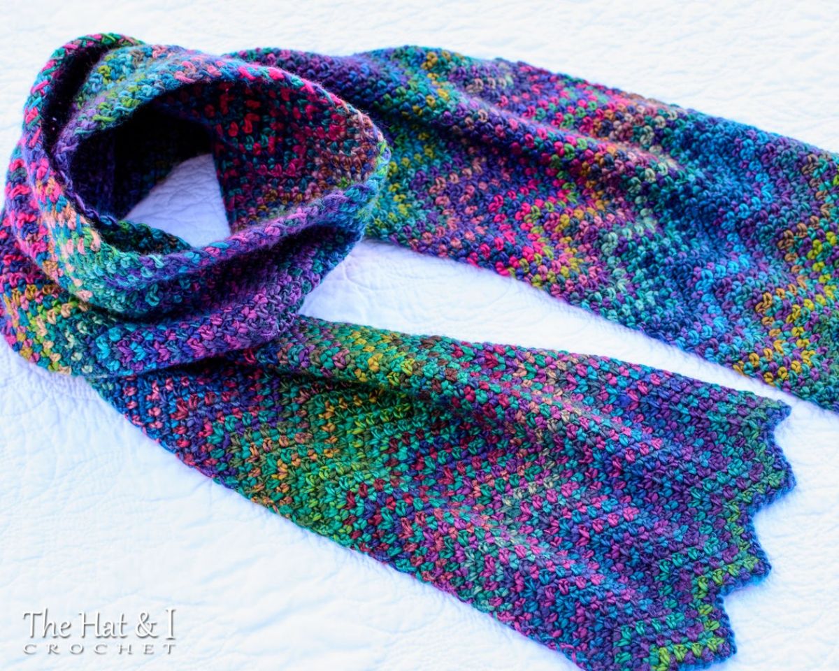 Purple, green, blue, pink, and yellow chevron style crochet scarf with a zig zag hem on a white fluffy blanket.