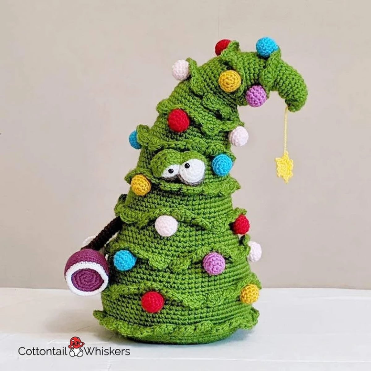 A large crochet green Christmas tree with drunk eyes in the middle and blue, pink, purple, and white balls all over.