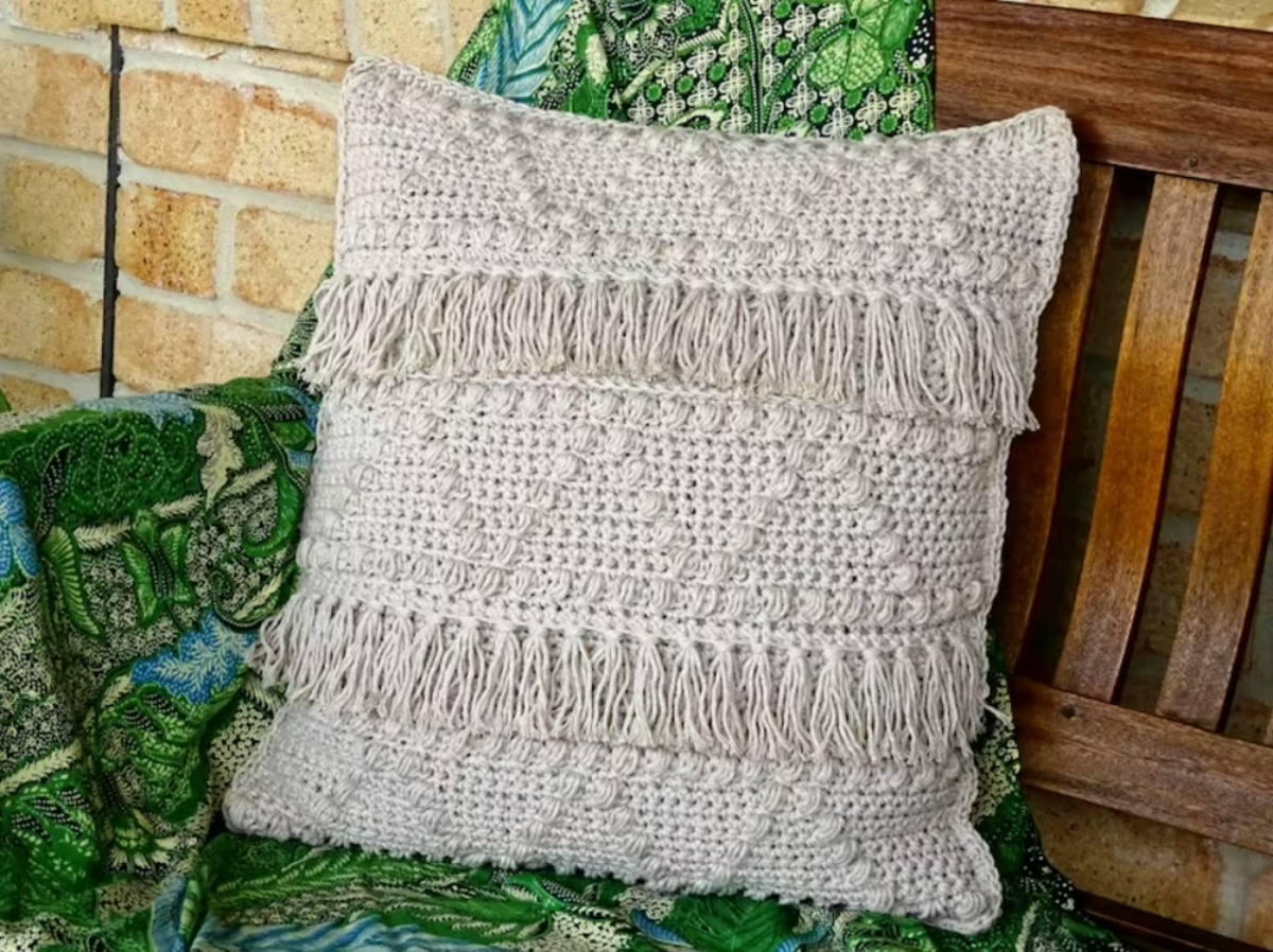 Cream crochet cushion with two rows of cream tassels and triangles with small bobbles stitched into the other rows on a wooden bench.