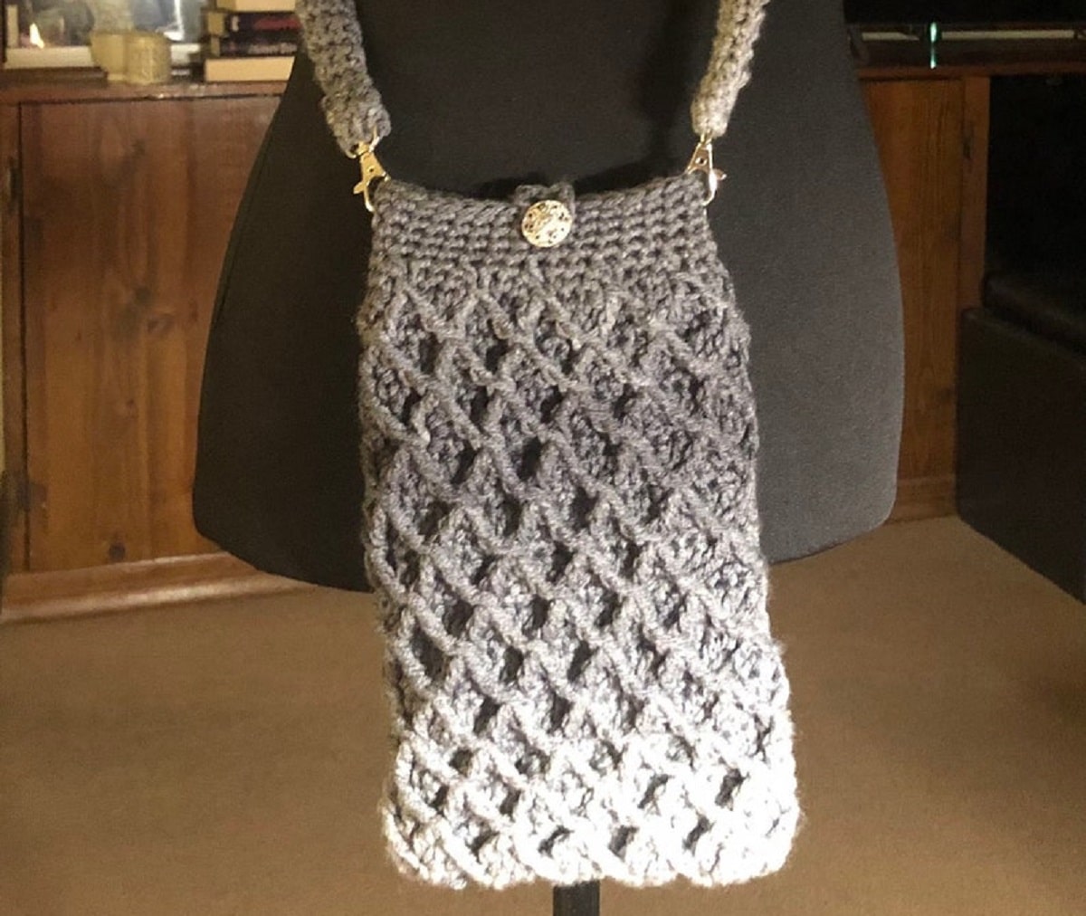 A gray and cream crochet bag with a diamond pattern stitching all over it, a white button at the top to fasten and a large strap. 