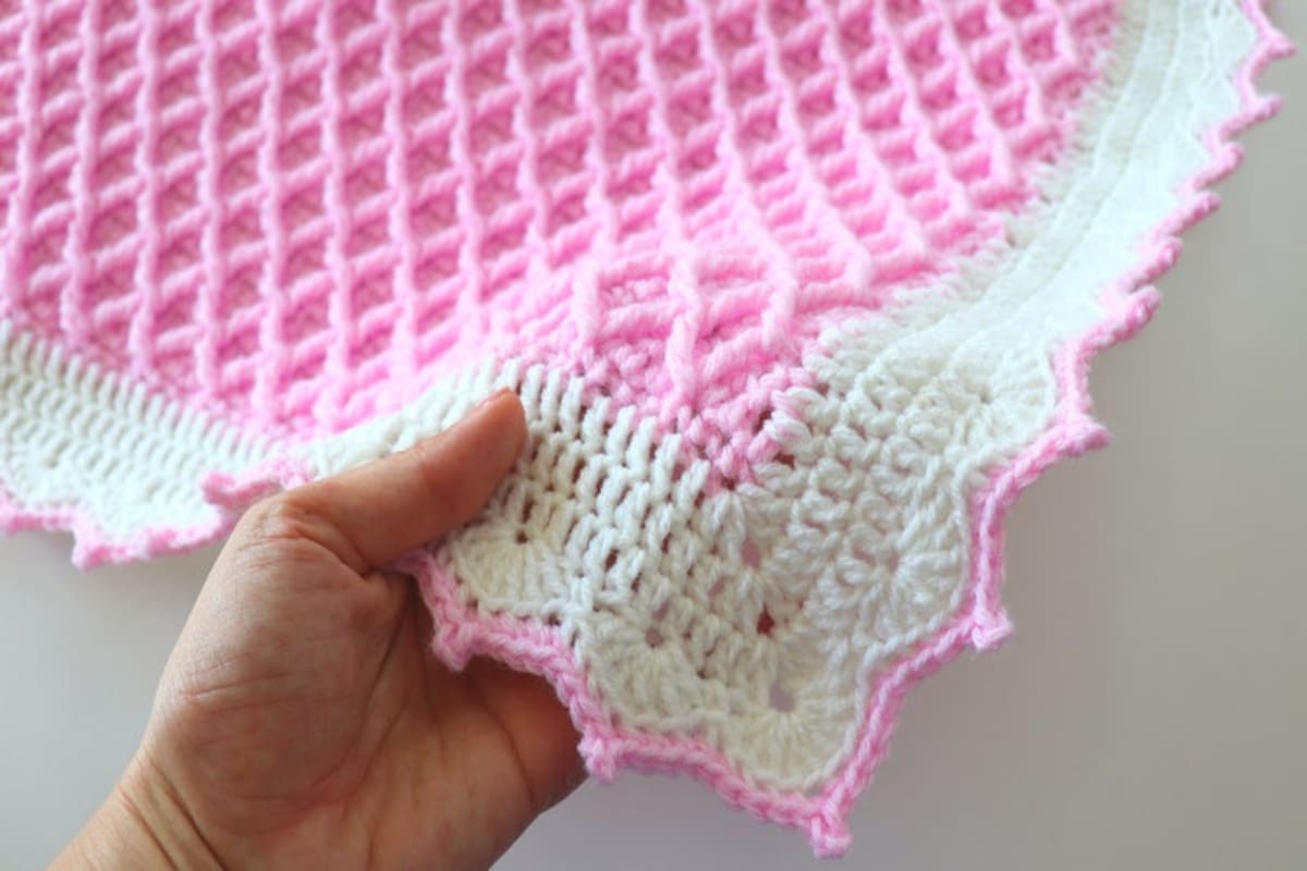 Corner of a pink waffle stitch crochet blanket with a white diamond shaped trim and narrow pink banding with small circles on it.
