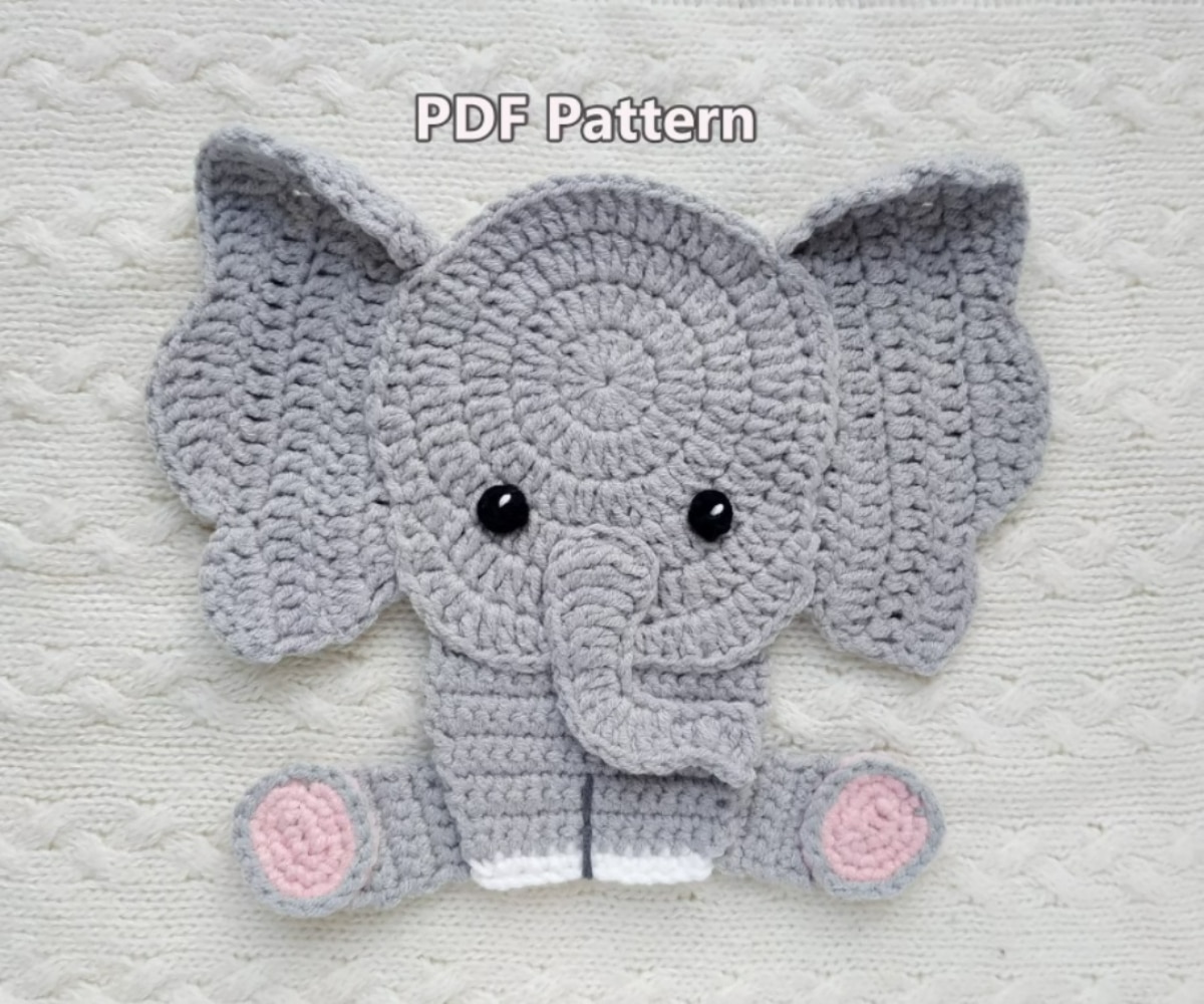 Small gray crochet elephant mat with large gray ears and small pink and white feet on a white blanket.