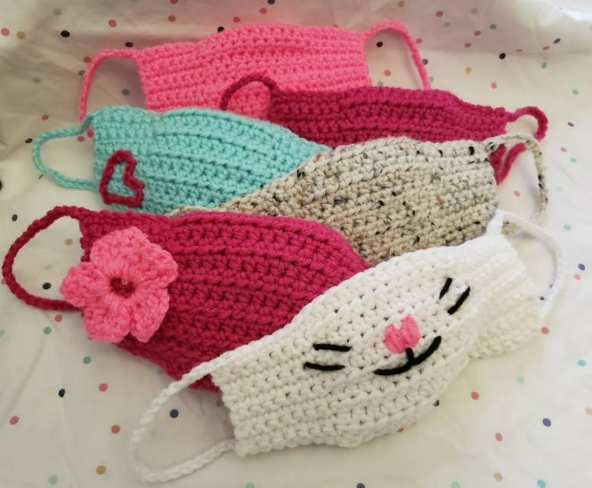 A small pile of pink, cream, red, and blue crochet facemasks sitting underneath a white facemask with a cat's face stitched on top in black.
