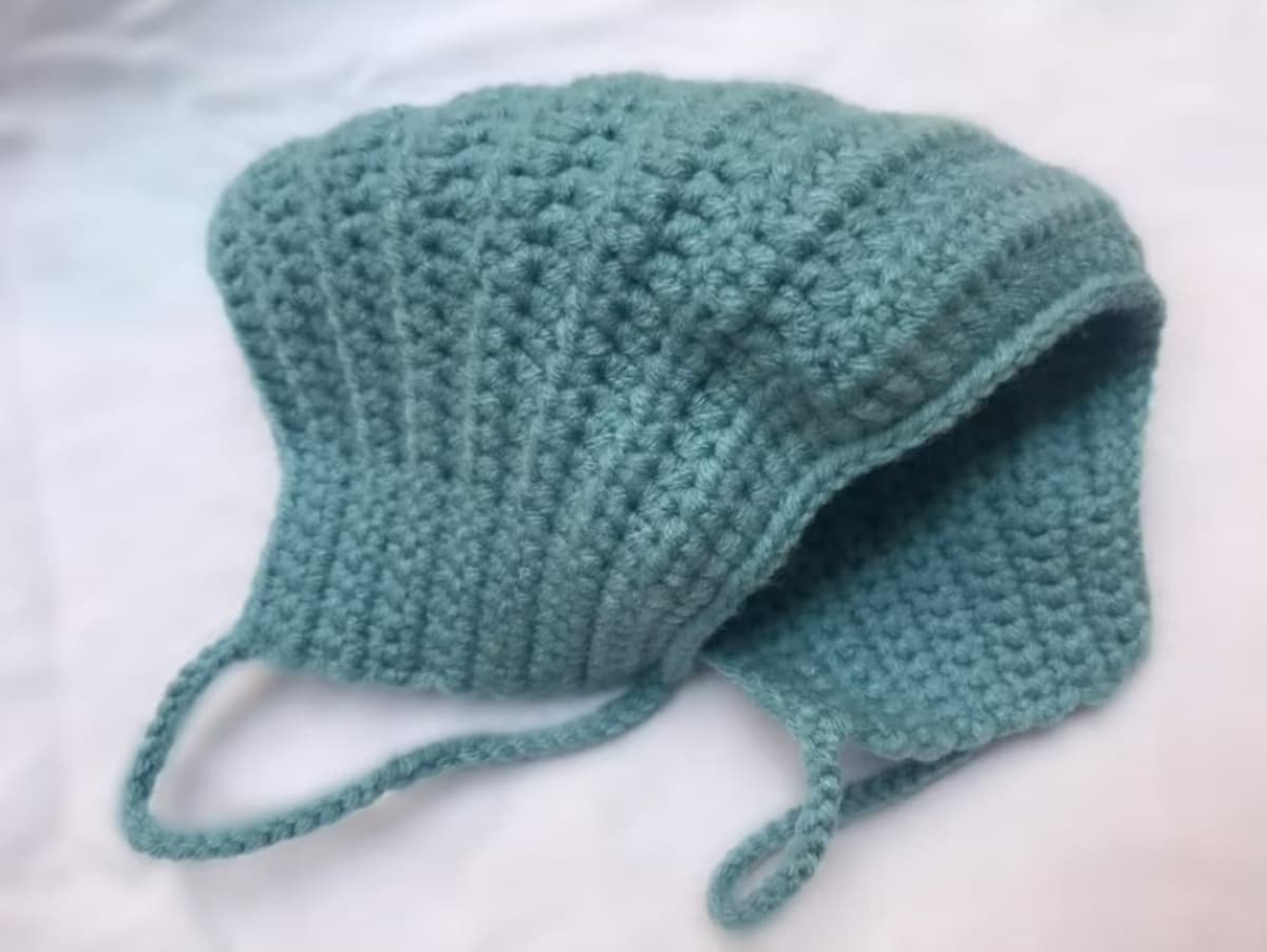 Teal crochet facemask with braided loops to secure over your ears folded in half on a white background.