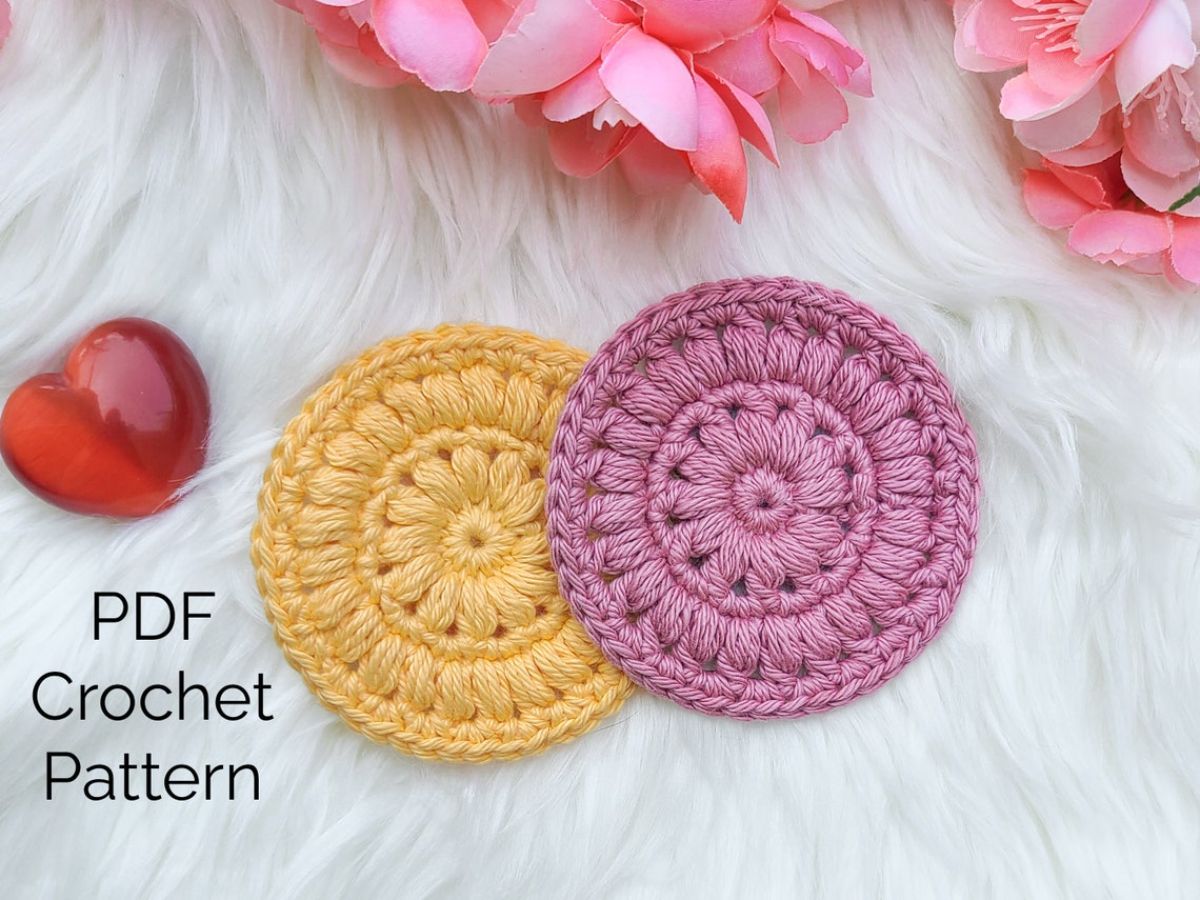 A yellow and pink crochet scrubbie next to each other with a small flower in the center on a white fluffy blanket.