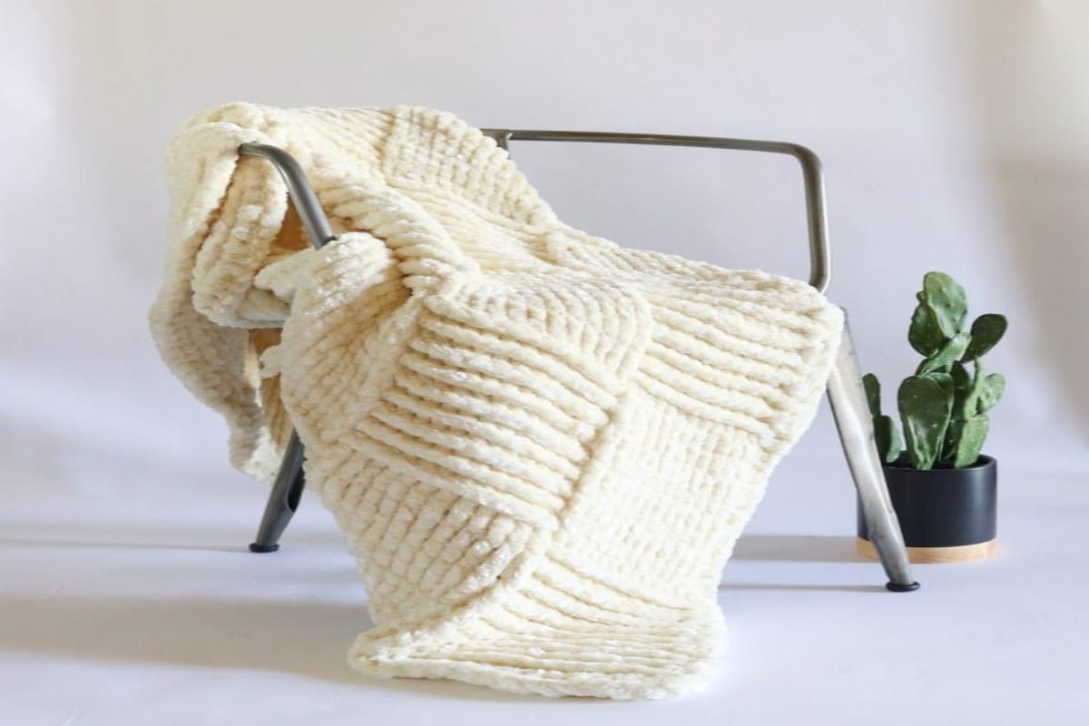 Large cream crochet blanket with a large basketweave pattern draped over a chair next to a small plant.
