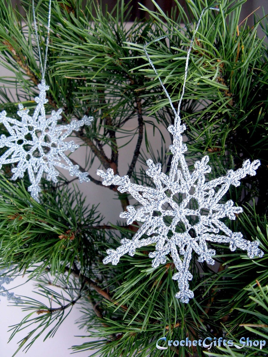 Two silver sparkling crochet snowflakes with ribbons attached hanging from a Christmas tree.