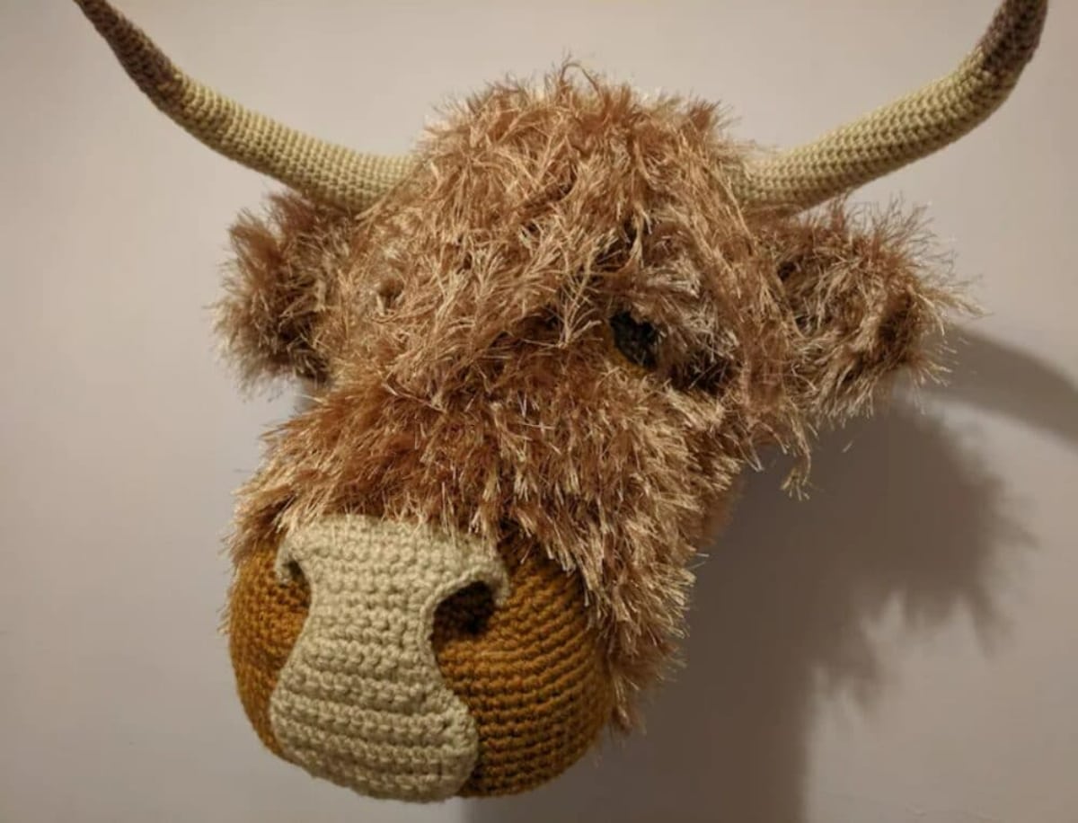 Brown crochet Highland Cow head with a white nose and horns mounted to a white wall.