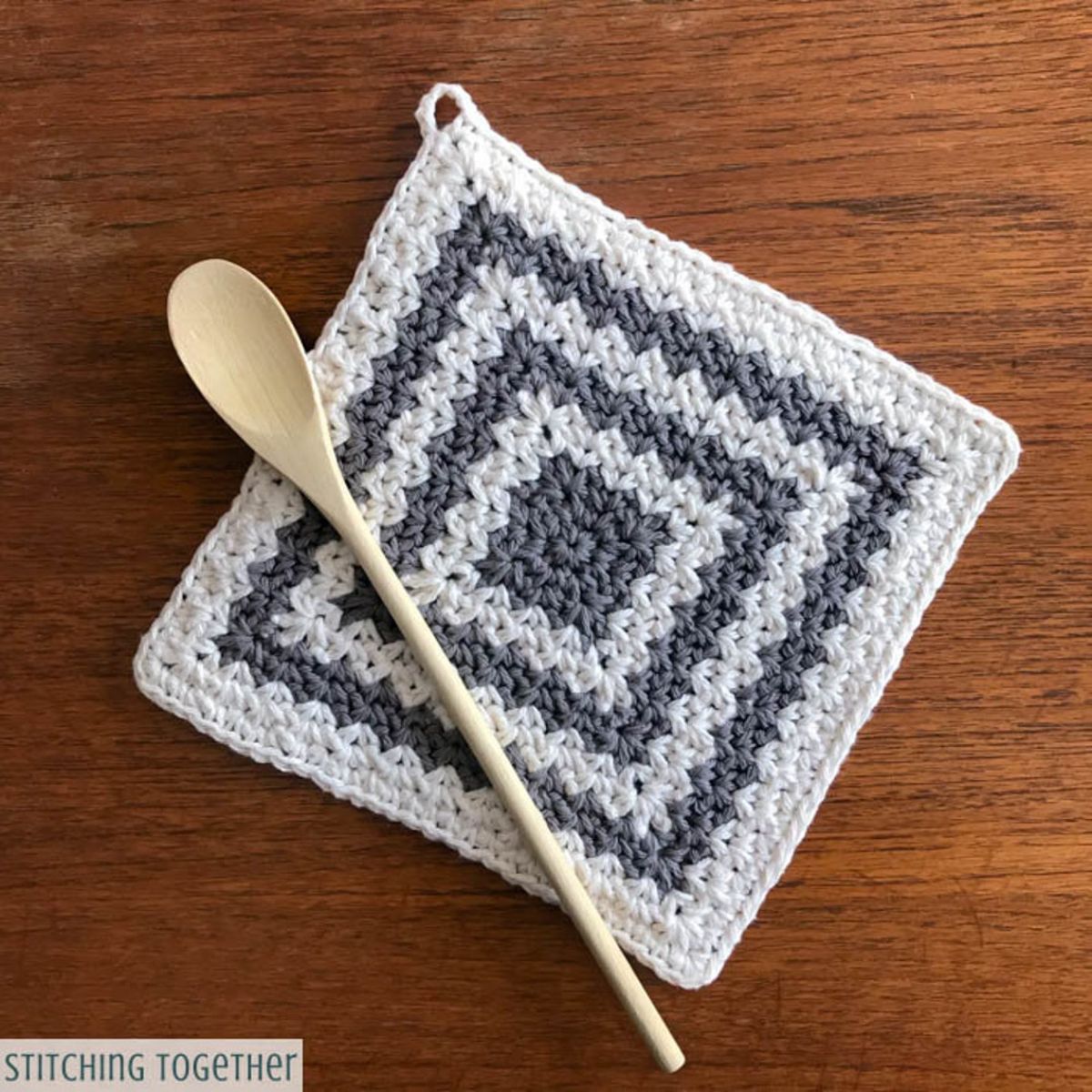 White and gray square crochet hot pad with gray squares getting smaller toward the center and a wooden spoon lying on top.