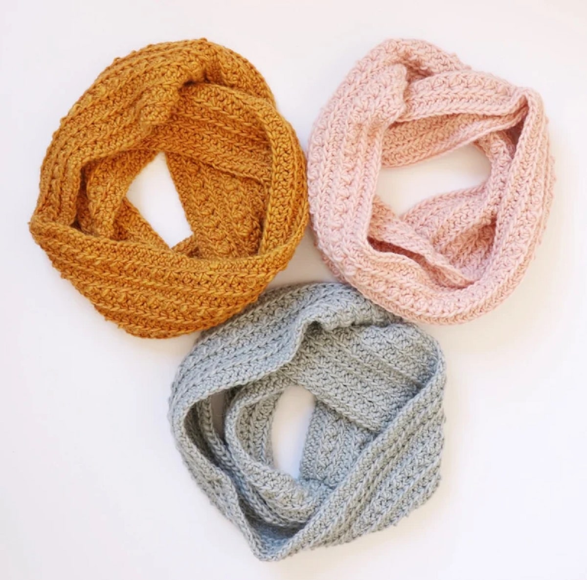 Mustard yellow light pink, and blue crochet cluster stitch infinity scarves in three circles on a white background.