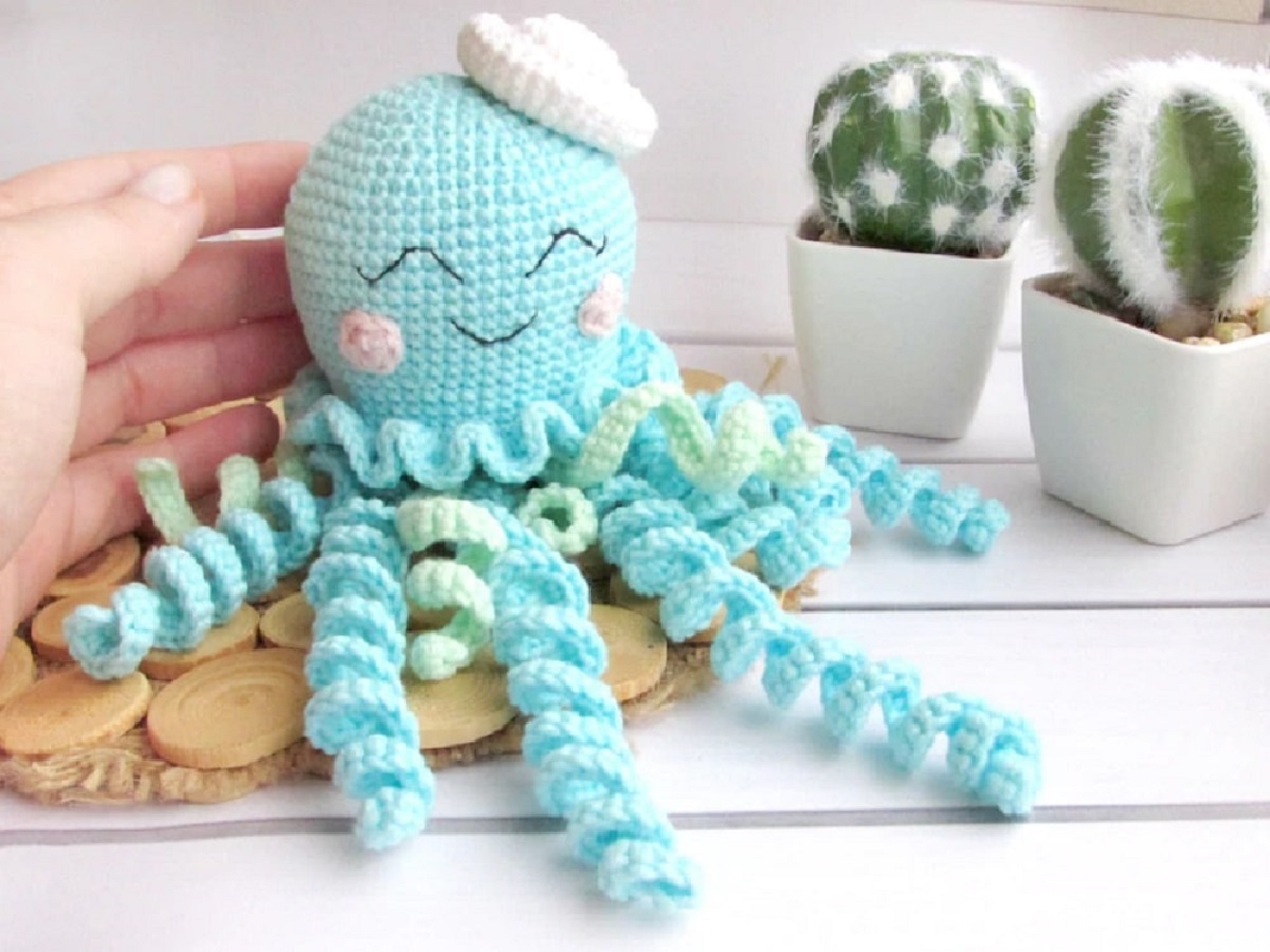 A small blue crochet jellyfish with a smiling face and pink small sailors cap on its head next to some small cacti. 