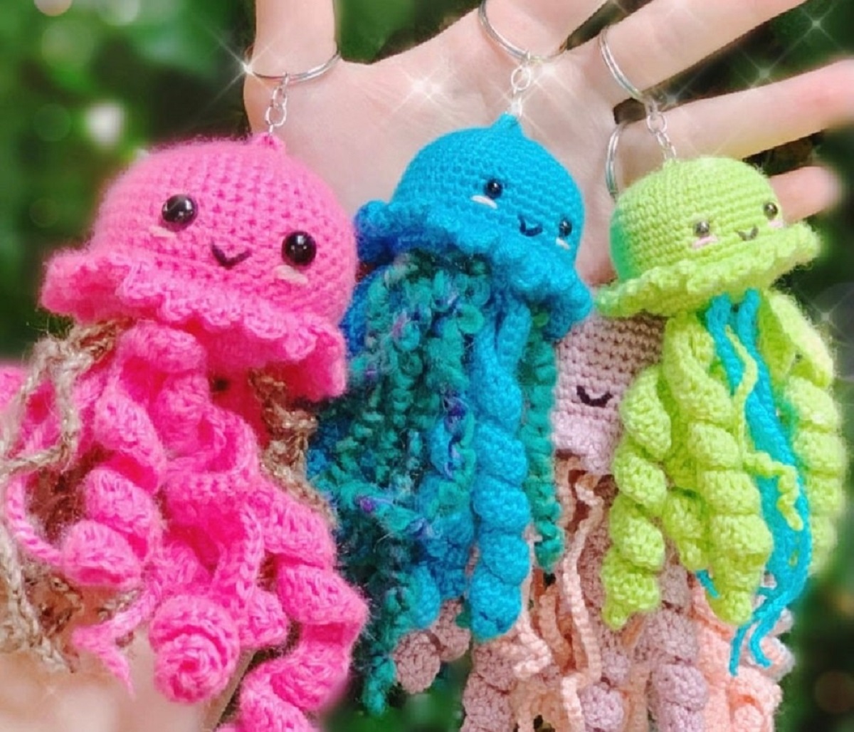 A hand holding three crochet jellyfish keychains in pink, blue, and yellow, with curled tentacles hanging from them.