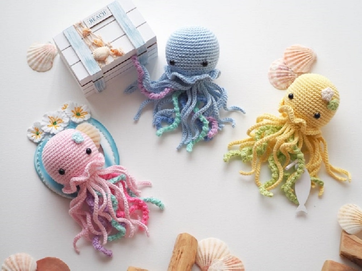 A pink, gray, and yellow crochet mini jellyfish lying on a white background with long pink, blue, yellow, and green tentacles.
