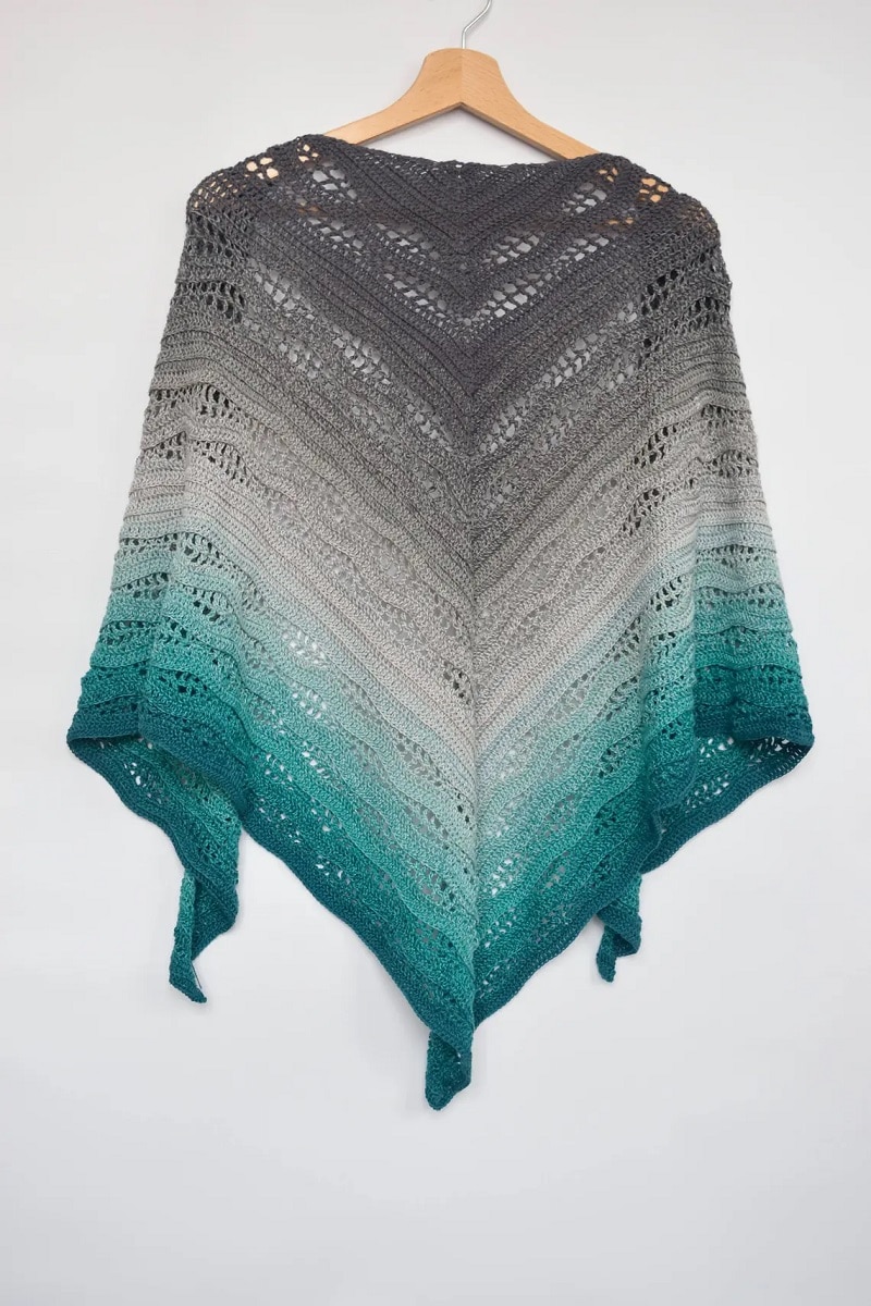 A gray, cream, and green ombre style crochet shawl hanging on a wooden hanger in front of a white background.