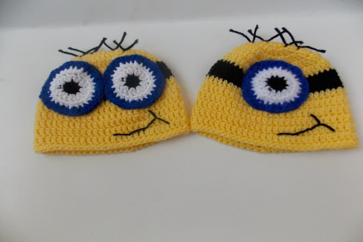 Two yellow crochet beanie hats, one with two white and black eyes, a smiling mouth, and spikes on top and the other has the same features but with one eye.