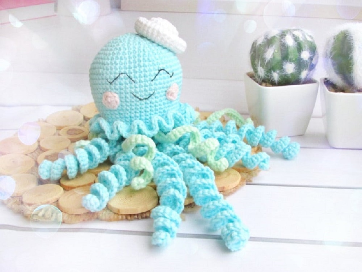 A blue crochet octopus with a small pink sailors hat on its head and blue and yellow curled tentacles spread around him.