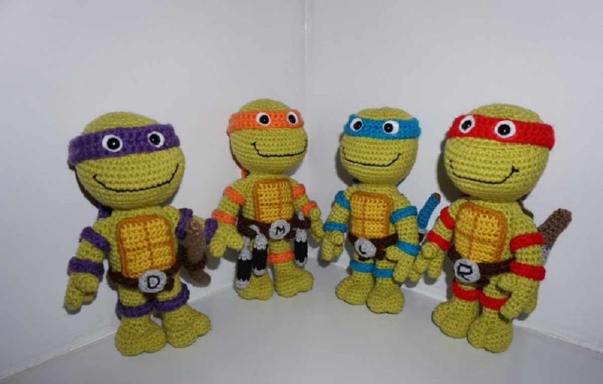 Four crochet teenage turtles with yellow chests, green bodies, and black smiling mouths. Each turtle has a different color eye mask on.