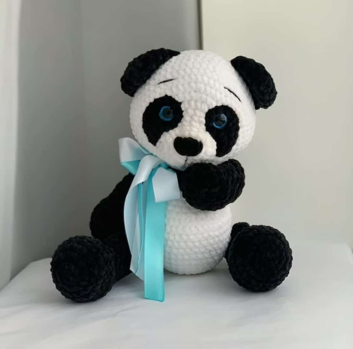 Stuffed crochet black and white panda bear on a white background with a silky blue and white bow around his neck.