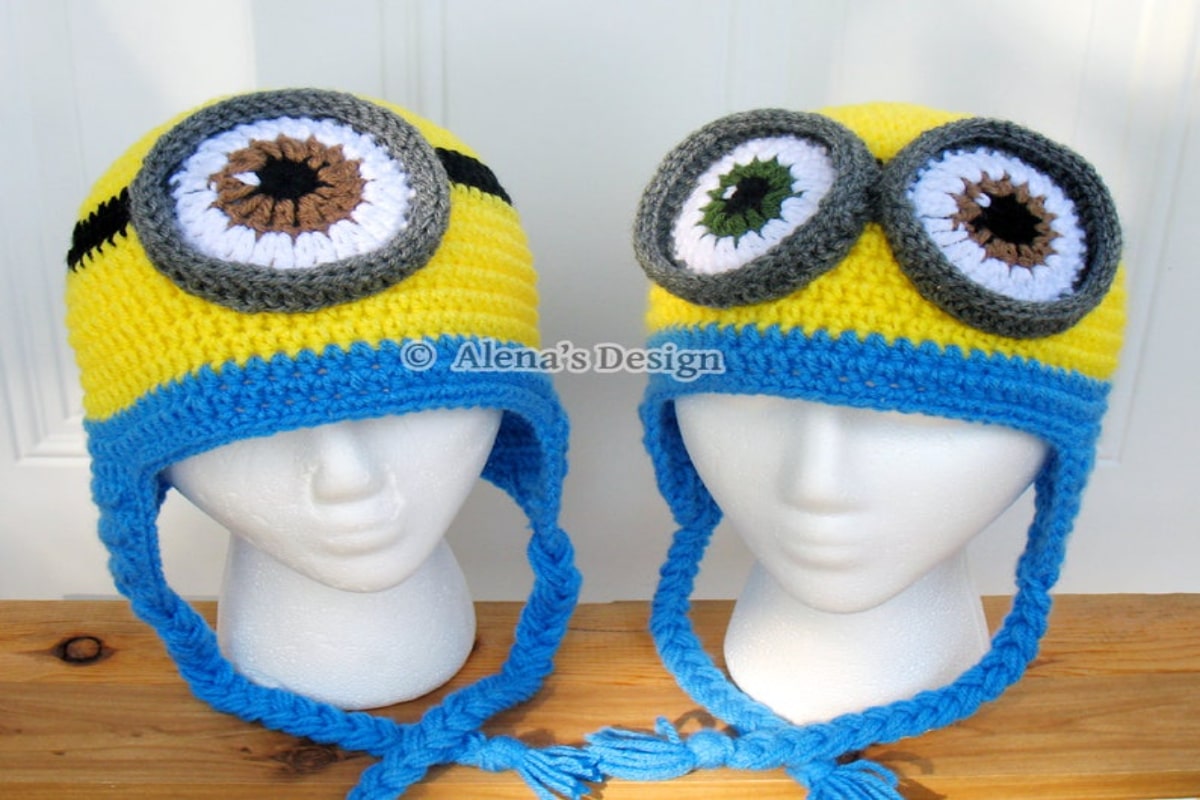  Two mannequins wearing yellow and blue crochet minion style hats one with one eye and the other has two eyes and blue braided ear flaps. 