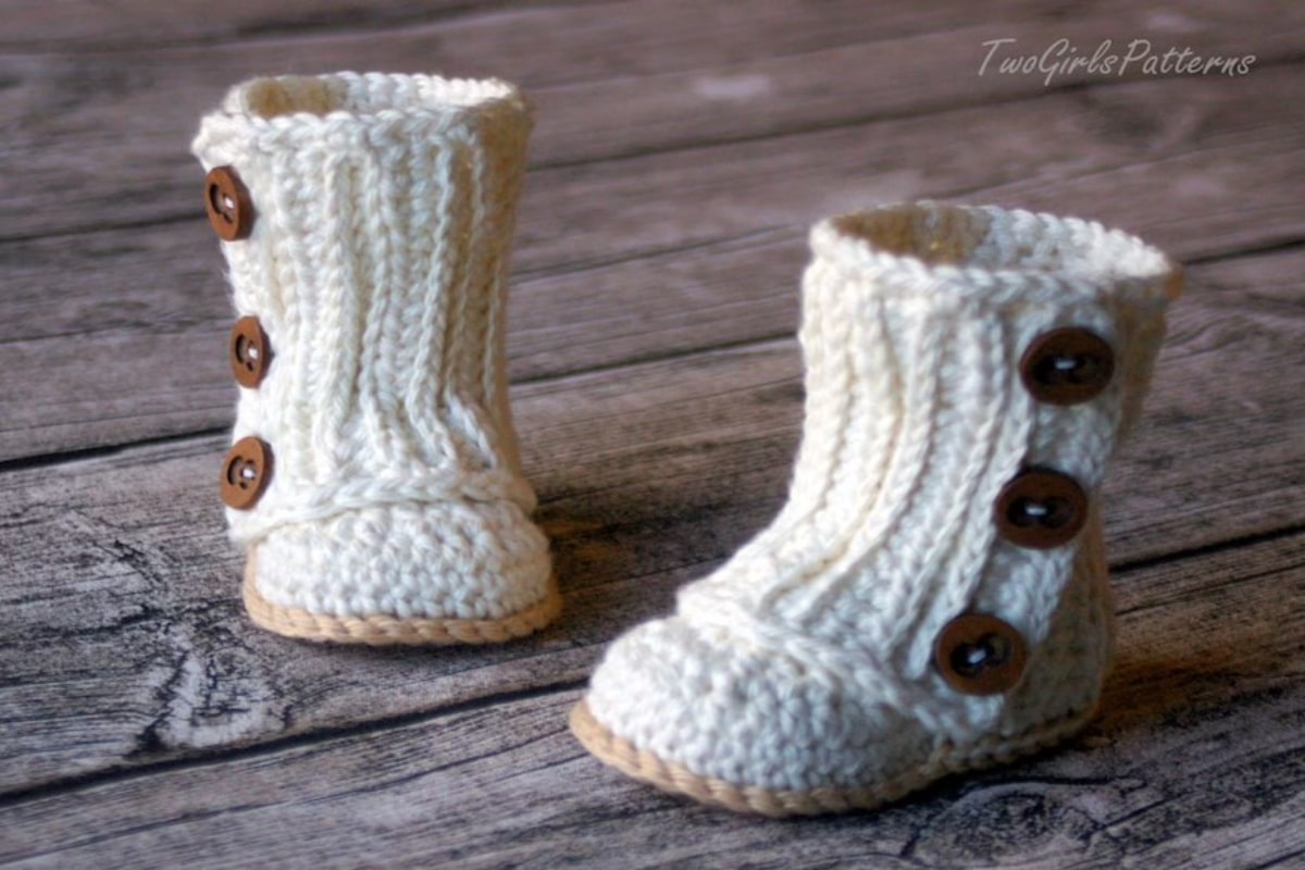 Calf length cream crochet baby booties with three buttons on the side to secure and a pale yellow sole on wooden flooring.