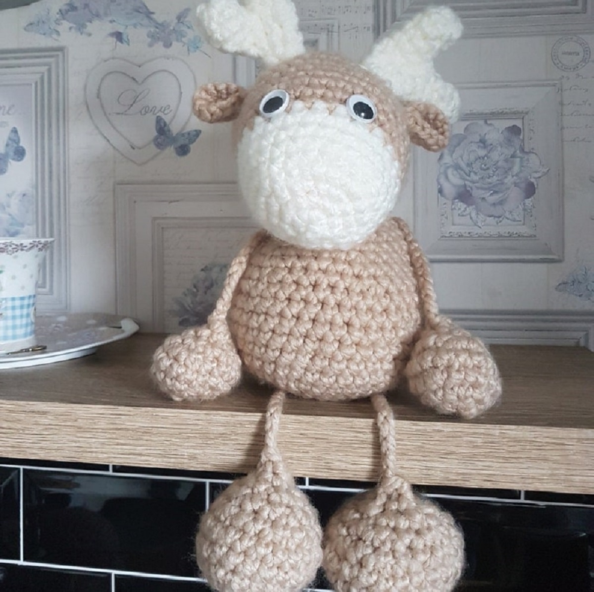 A large brown crochet reindeer with a white nose and antlers and oversized feet sitting on a wooden shelf.