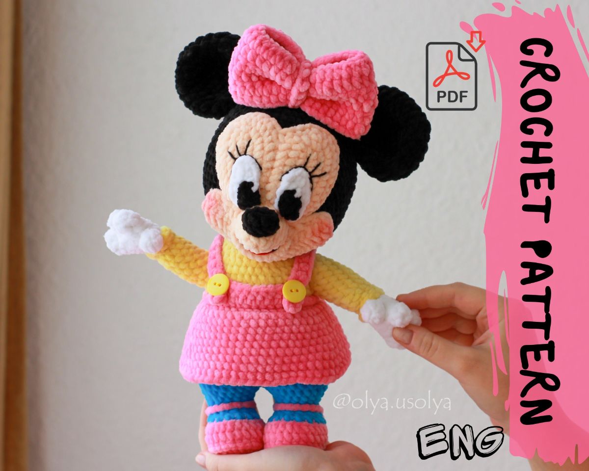 Small and squat baby-style crochet Minnie Mouse in a yellow top and pink dress with a pink bow and shoes being held up by some hands.
